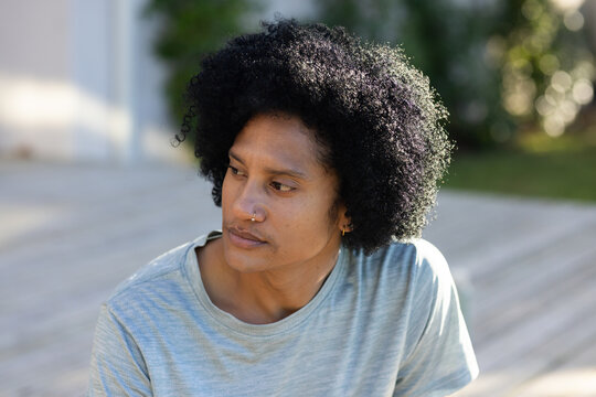African American man sitting on wood deck wearing grey tee, nose ring, hoop earring, blurred shrubs