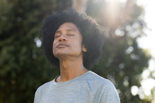 African American man standing eyes closed, tilting upward in park, in gray tee, nose ring, earring