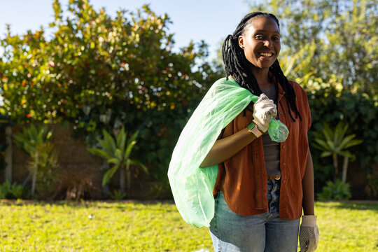 Adult African woman standing slinging green bag over shoulder wearing gloves in yard, copy space