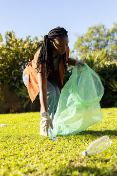 African American woman gloved crouching on sunlit lawn in rust shirt picking bottles into green bag