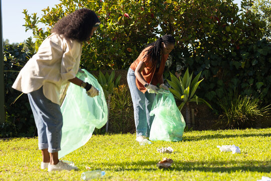African women collecting litter on lawn by hedge, holding green bags and wearing gloves