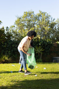 African American male bending and picking up litter wearing gloves with green trash bag on lawn
