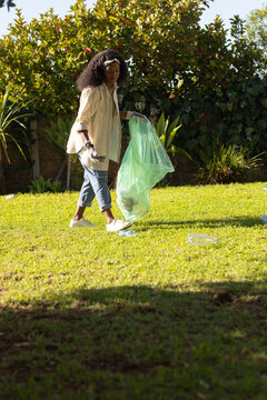 Woman walking across grassy yard collecting litter near bottles with green trash bag wearing gloves