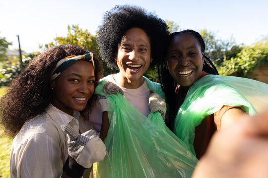 African American friends taking phone selfie in sunny backyard holding green trash bags and gloves