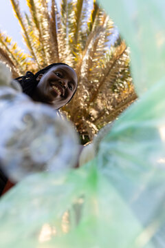 African woman with braided hair smiling, leaning over green plastic bag at park holding bottle cap