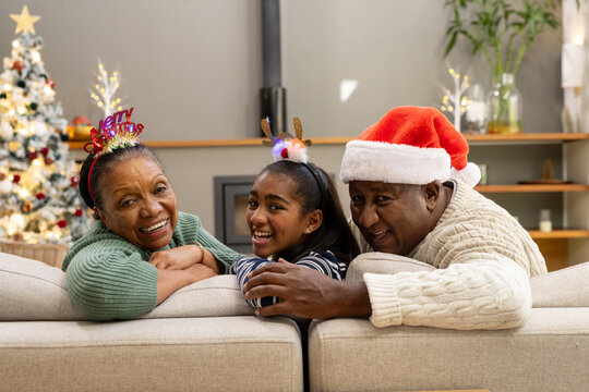 African American family senior and youth leaning over sofa at home, Christmas tree lit wearing hats