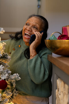 Senior African American woman in green knit sweater holding phone, laughing at home by flocked tree