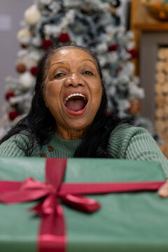 Senior African American woman holding wrapped green gift in sweater at home before decorated tree