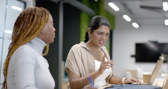 Diverse female team pausing typing, working, gesturing toward laptop, notes, coffee at study table
