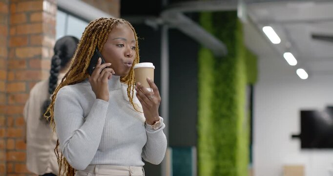 African American woman receiving call, holding smartphone and cup, testing sip, smiling in office