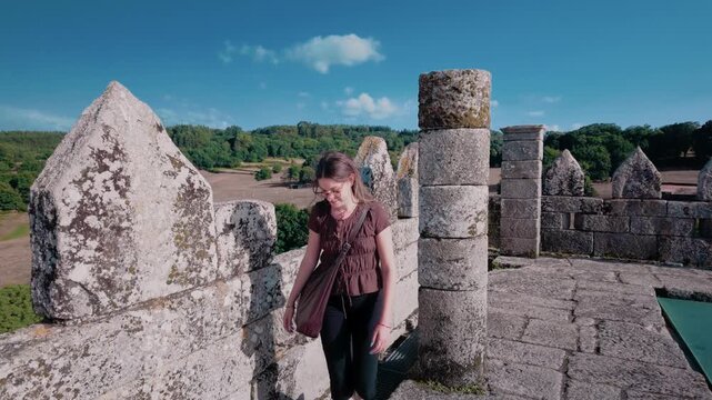 Young thoughtful woman with long hair walking on the ramparts of a medieval fortress, appreciating the historical architecture and surrounding verdant scenery on a beautiful sunny day