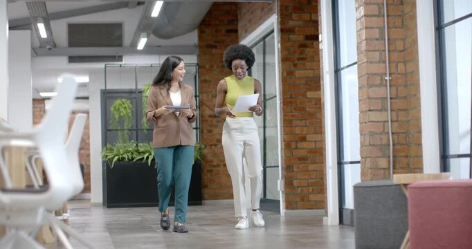 Two women coworkers walking down hall left holding tablet right reading pages laughing about work