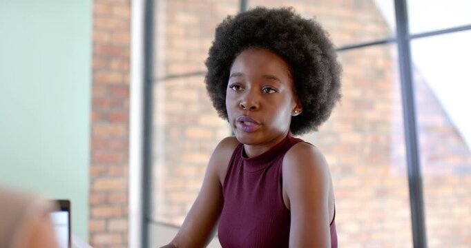 African woman in maroon sitting at office laptop with docs reacting to colleague comment laughing