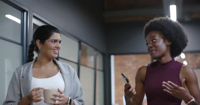 Diverse women starting chat walking glass office corridor showing phone holding mug laughing