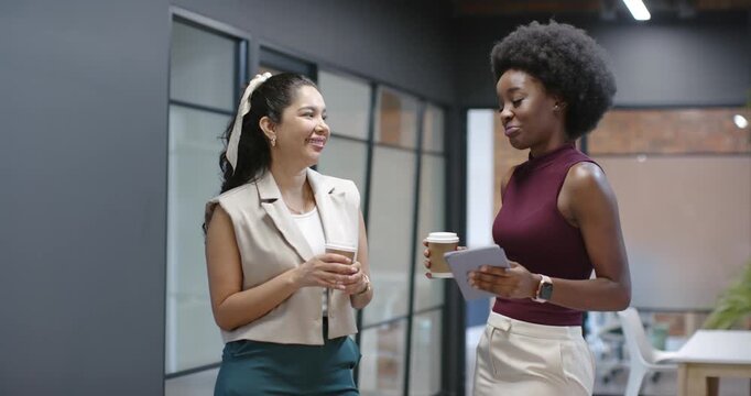 Diverse female coworkers talking in office hall after tablet glance holding coffees and smartwatch