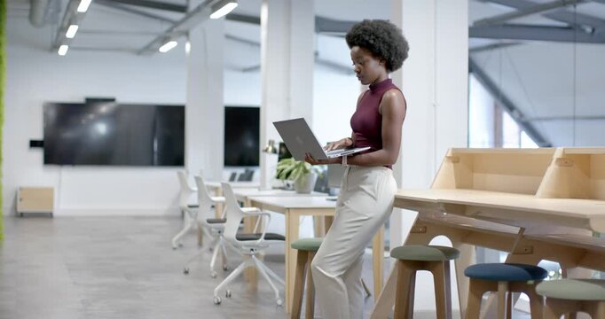 African American woman holding laptop, typing, editing report by high table in office, copy space