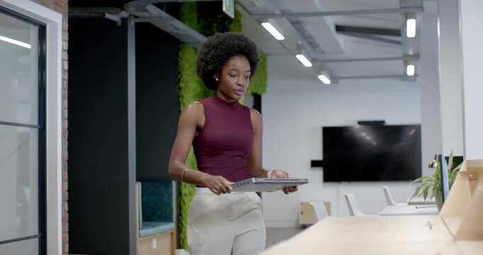 African American woman walking across office carrying laptop, preparing bench for work, typing