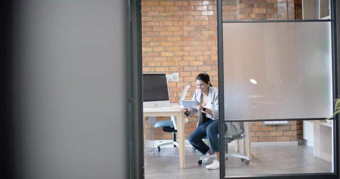 Adult Indian woman in blazer reading papers in glass office comparing on tablet walking down hall