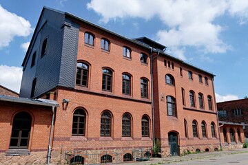Fototapeta premium An old renovated industrial red brick building featuring multiple windows and a unique slate shingle accent wall against a bright blue cloudy sky.