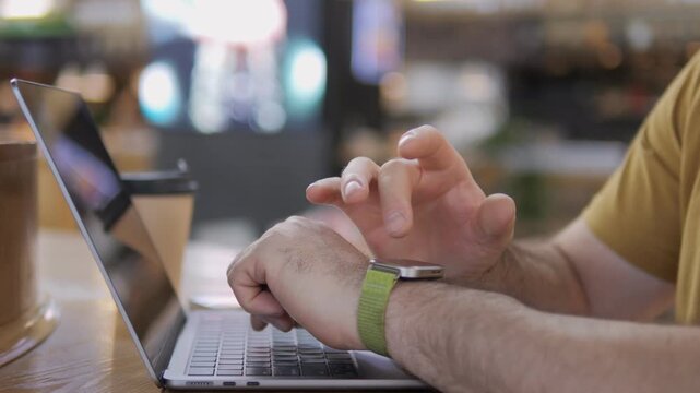 Close-up of hands resting on laptop in cafe. Person wears green wristband, yellow shirt. Fingers gently tap, suggest idle or thoughtful moment. Background softly blurred with warm lights