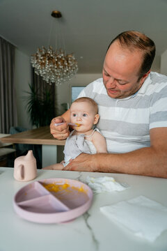Vertical full shot of a father sitting at a modern kitchen table, feeding his baby with a spoon. Ideal for social media stories, vertical ads, family lifestyle, and parenting blogs