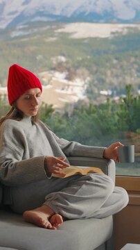 A woman reading by a large window and enjoying scenic snowy mountain view.