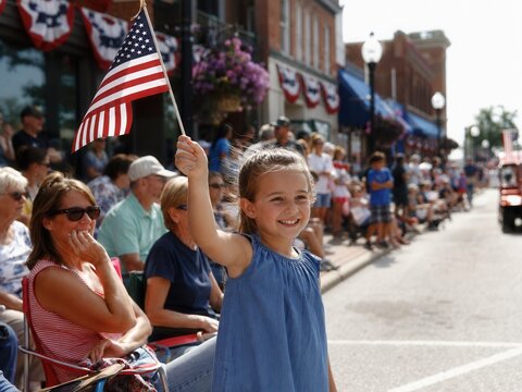 L&auml;chelndes M&auml;dchen schwenkt bei einer Labor Day Parade in einer amerikanischen Kleinstadt die US-Flagge, w&auml;hrend Zuschauer entlang der festlich geschm&uuml;ckten Stra&szlig;e warten.