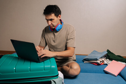 Portrait of traveler male preparing for travel, working on his laptop placed on a teal luggage, with neatly folded clothes stacked beside him on a couch, organizing logistics for his vacation.