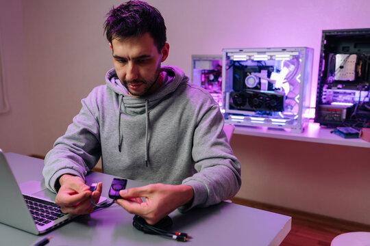 Man in hoodie focusing on connecting audio cables and an adapter, sitting in front of laptop with illuminated gaming pc rigs visible in background.