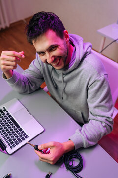 Young creative professional sitting at desk with laptop. He holding audio cable and smiling mischievously at camera. Scene lit by purple neon light, creating modern techy atmosphere.