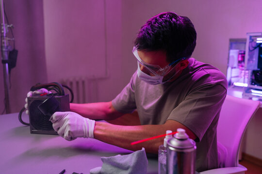 Technician wearing safety goggles, mask gloves cleaning computer power supply unit with compressed air, ensuring computer hardware maintenance and proper functioning.