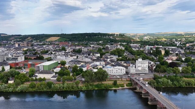 Zeitraffer (Timelapse)  Drohnenaufnahme von Trier mit der antiken R&ouml;merbr&uuml;cke &uuml;ber die Mosel, umgeben von Stadtarchitektur und gr&uuml;nen H&uuml;geln unter bew&ouml;lktem Himmel an einem Sommertag.