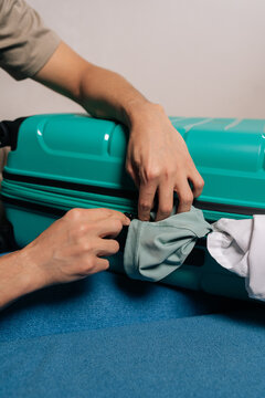 Close-up hand of traveler struggling to zip an overpacked turquoise suitcase, pushing clothes inside while preparing for a journey, symbolizing common packing challenges and travel readiness.