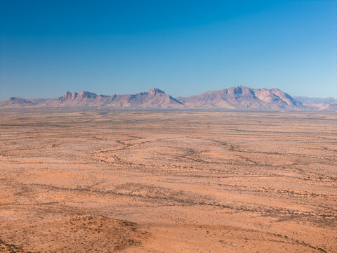 An aerial shot shows the vast desert landscape and distant rocky mountains under a clear sky in Spitzkoppe, Namibia.