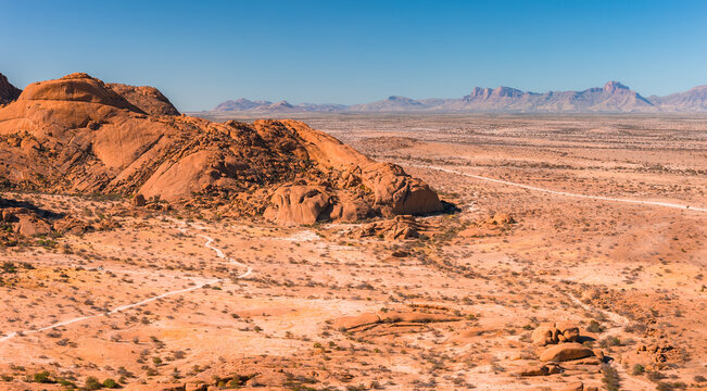 An aerial perspective shows the vast desert landscape of Spitzkoppe in Namibia, featuring large granite rock formations, dry plains, and distant mountains.