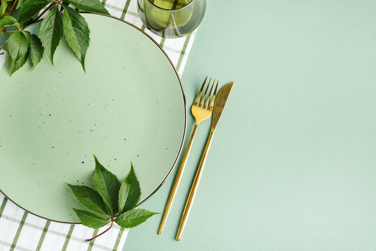 Cleanly set table with green plate and golden cutlery decorated with fresh green leaves and gingham napkin for festive occasion