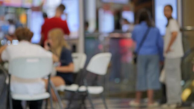 Blurred indoor event space with people moving. Woman walks away, wearing gray shirt and jeans. Others sit around tables or stand near screens. Neutral expressions suggest casual activity
