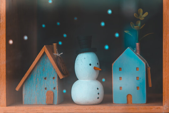 Cute wooden snowman figurine wearing a top hat stands between miniature blue houses against a cozy winter background, Charming Christmas holiday decoration concept, Perfect for festive seasonal themes