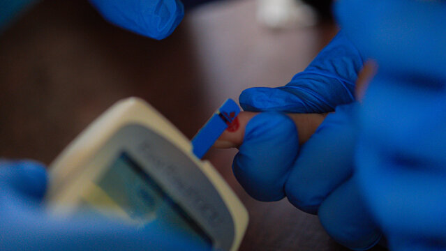 View of a medical professional wearing blue gloves performing a blood glucose test on a patient's finger in Southwest Papua, Indonesia.