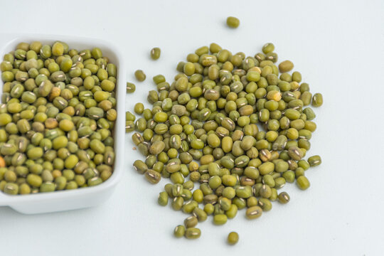 Raw green mung beans in a small white ceramic bowl next to a scattered pile
