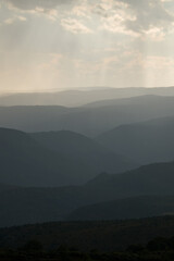 Obraz premium Dramatic mountain landscape with sun rays shining through storm clouds and distant rain over layered hills.