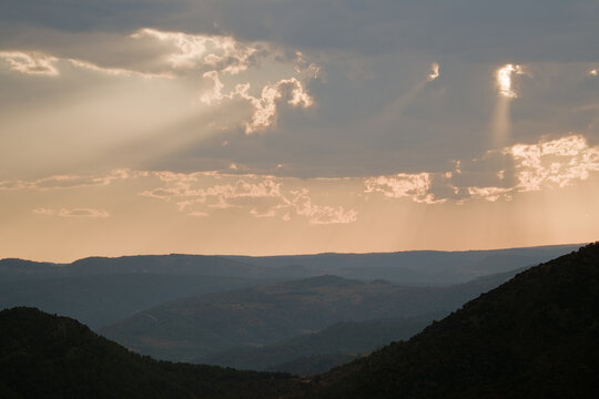 Dramatic sunset over layered mountain hills with golden sun rays shining through dark clouds.