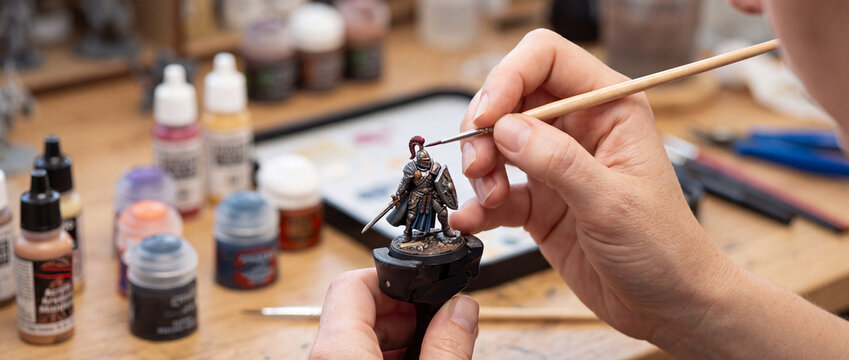 Hands painting a fantasy miniature figure at a hobby workbench with paints and brushes