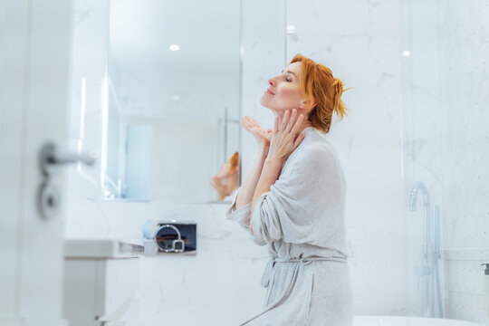 Middle aged woman doing face building yoga facial gymnastics self massage and rejuvenating exercises looking in bathroom mirror. Natural procedures for skin care. Home Beauty treatment routine