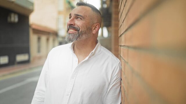Man leans shoulder on brick wall, looks up smiling with beard and shaved sides, wearing white shirt on a street lined with buildings; quiet contentment.