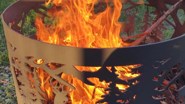 Bright flames blaze inside carved steel bowl, creating warm outdoor campfire in green backyard. Gentle smoke rises from outdoor campfire as wood burns brightly during cozy summer evening.