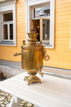 Antique brass tea samovar standing on a white wooden table near a yellow house wall. Ideal stock photo for Russian culture, traditional tea brewing, antiques, and heritage.