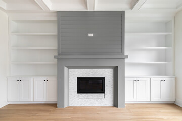 Fototapeta premium A fireplace detail with white built-in shelving and cabinets, white oak flooring, white coffered ceiling, and a grey accent surround and mantel.