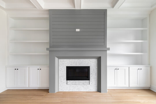 A fireplace detail with white built-in shelving and cabinets, white oak flooring, white coffered ceiling, and a grey accent surround and mantel.