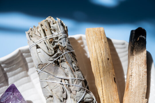 close up bundle of dried sage with palo santo sticks and amethyst crystal on a ceramic dish